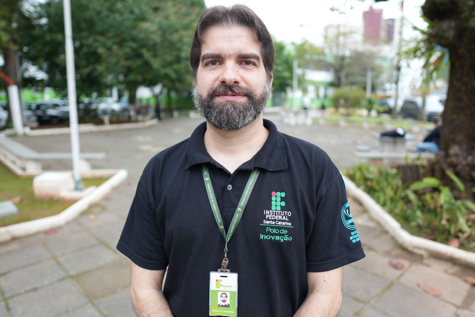 A imagem mostra  o diretor do Polo de Inovação do IFSC, Edison Antonio Cardoso Aranha Neto, um homem em pé em área externa arborizada, em frente a um pátio com bancos e árvores. Ele olha para a câmera e sorri levemente. Tem pele clara, cabelo castanho escuro penteado para trás e barba cheia. Veste camiseta polo preta com o logotipo do Instituto Federal de Santa Catarina (IFSC) bordado, acompanhado dos dizeres “Polo de Inovação”. No pescoço, usa um cordão verde com crachá de identificação institucional. O fundo está desfocado, mas é possível ver árvores, parte de um prédio e alguns carros estacionados.