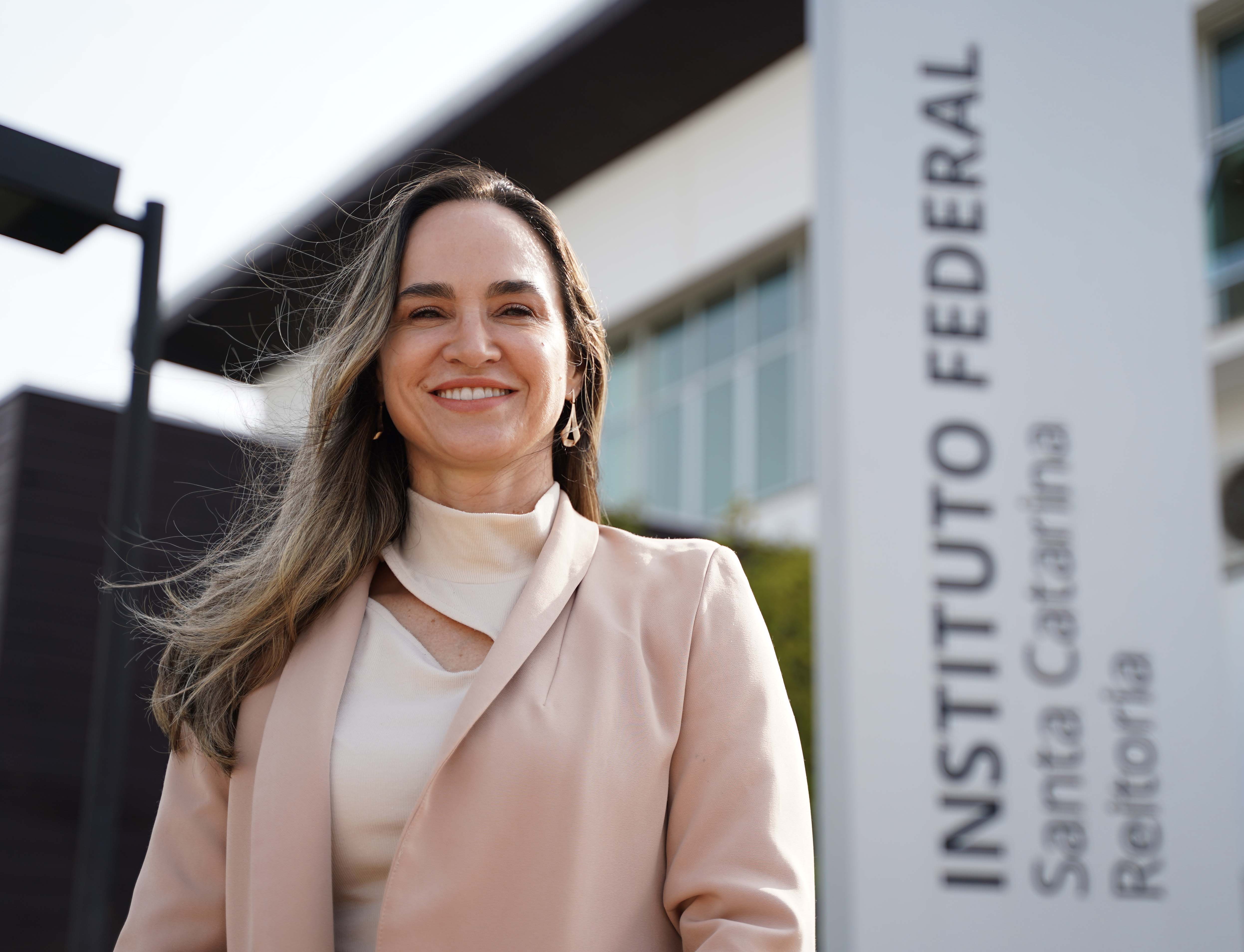Foto da pró-reitora de Ensino do IFSC, Eliana Cristina Bär. Mulher de pele clara e cabelos castanhos com mechas loiras, soltos e levemente ondulados, aparece sorridente em primeiro plano. Ela veste uma blusa clara de gola alta e um blazer em tom rosado. Ao fundo, desfocado, vê-se parte da fachada de um prédio moderno, com janelas de vidro e um totem vertical onde se lê: “INSTITUTO FEDERAL Santa Catarina Reitoria”. A foto foi tirada em ambiente externo, com luz natural destacando o rosto da mulher.